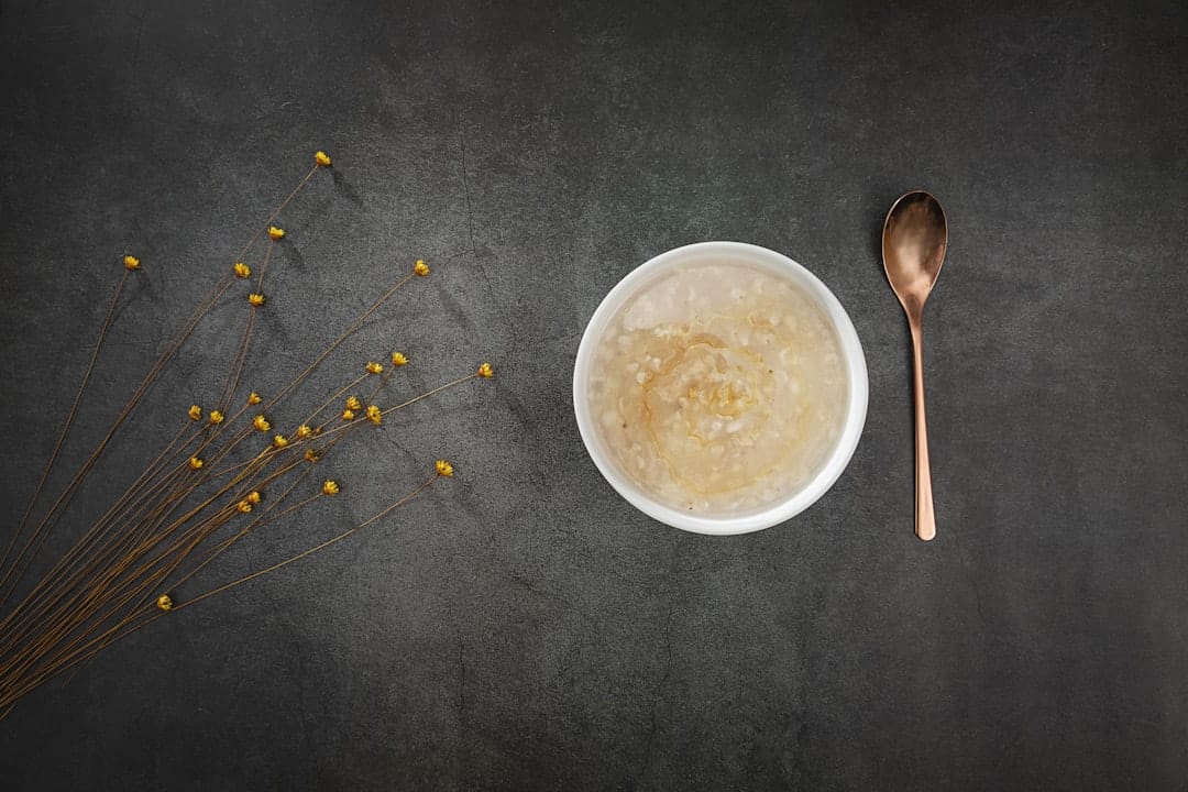 White bowl of oat porridge drizzled with honey beside a copper spoon and dried yellow flowers