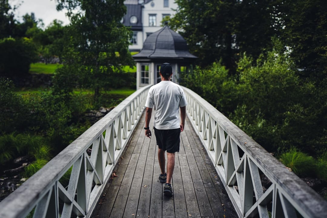 Man in a white T-shirt walking across a wooden footbridge toward a gazebo in a green park