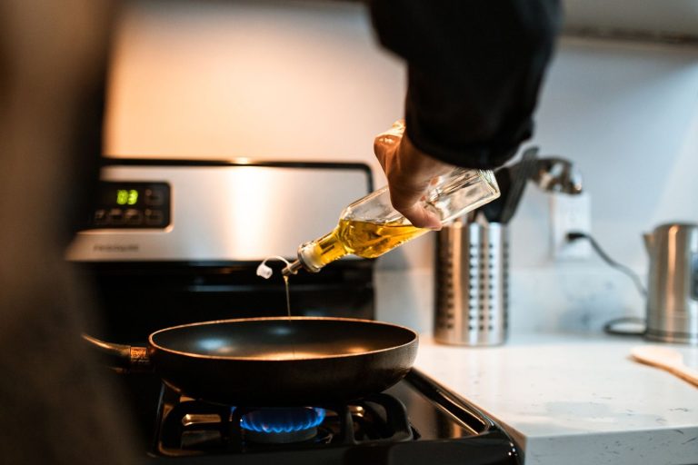 Golden cooking oil being poured from a glass cruet into a frying pan on a gas hob