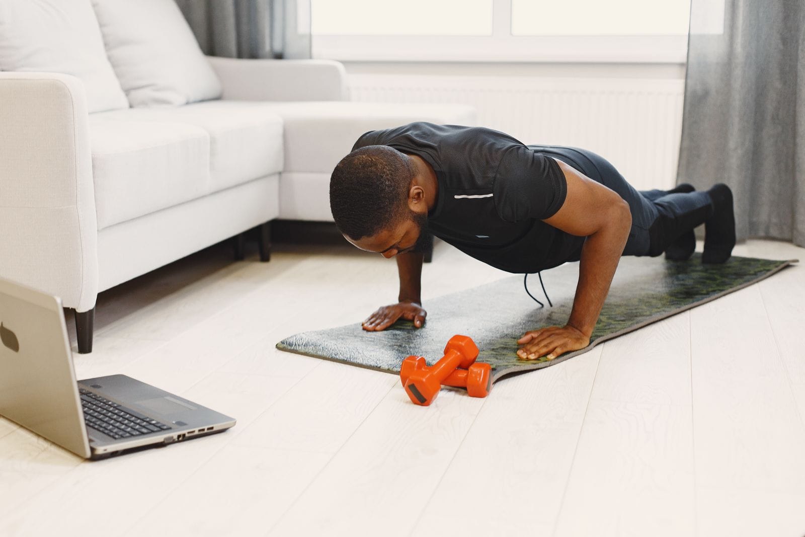 Man doing a push-up on a mat in a living room with small dumbbells and a laptop beside him