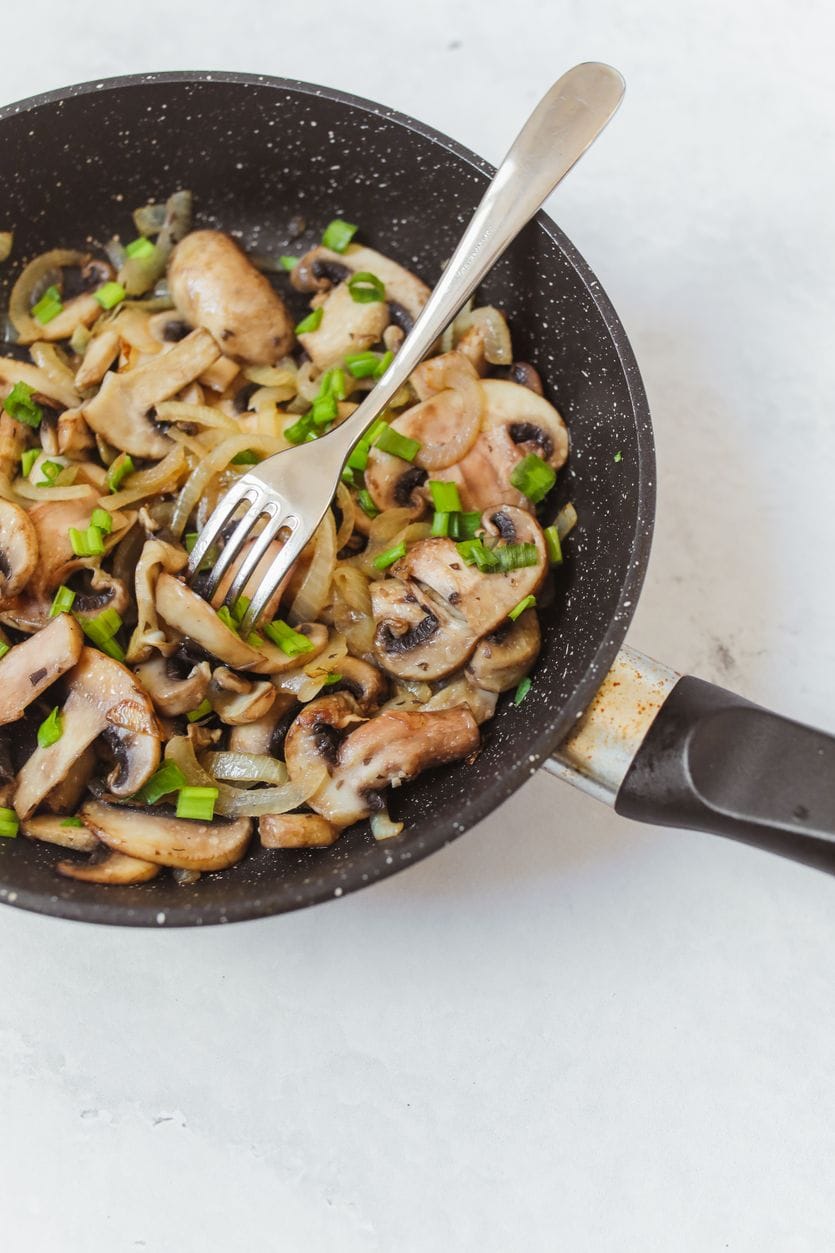 Sliced button mushrooms sauteed with onion and topped with chopped spring onion in a dark frying pan with a fork