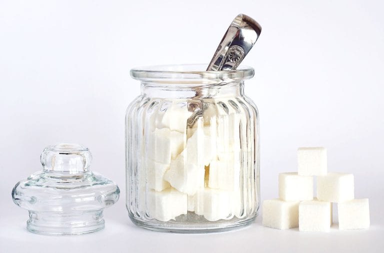 Glass jar of white sugar cubes with a silver spoon, lid beside it and a stack of cubes on a white background