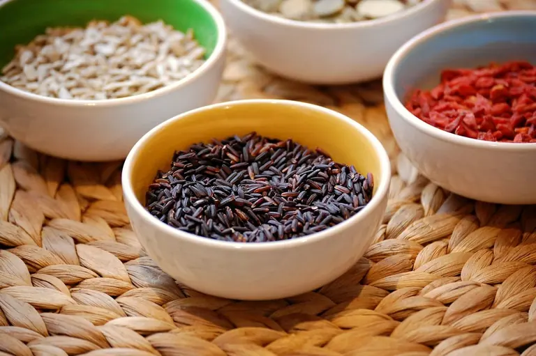 Bowls of dark purple-black rice grains on a woven mat, with bowls of sunflower seeds, pumpkin seeds and goji berries around it