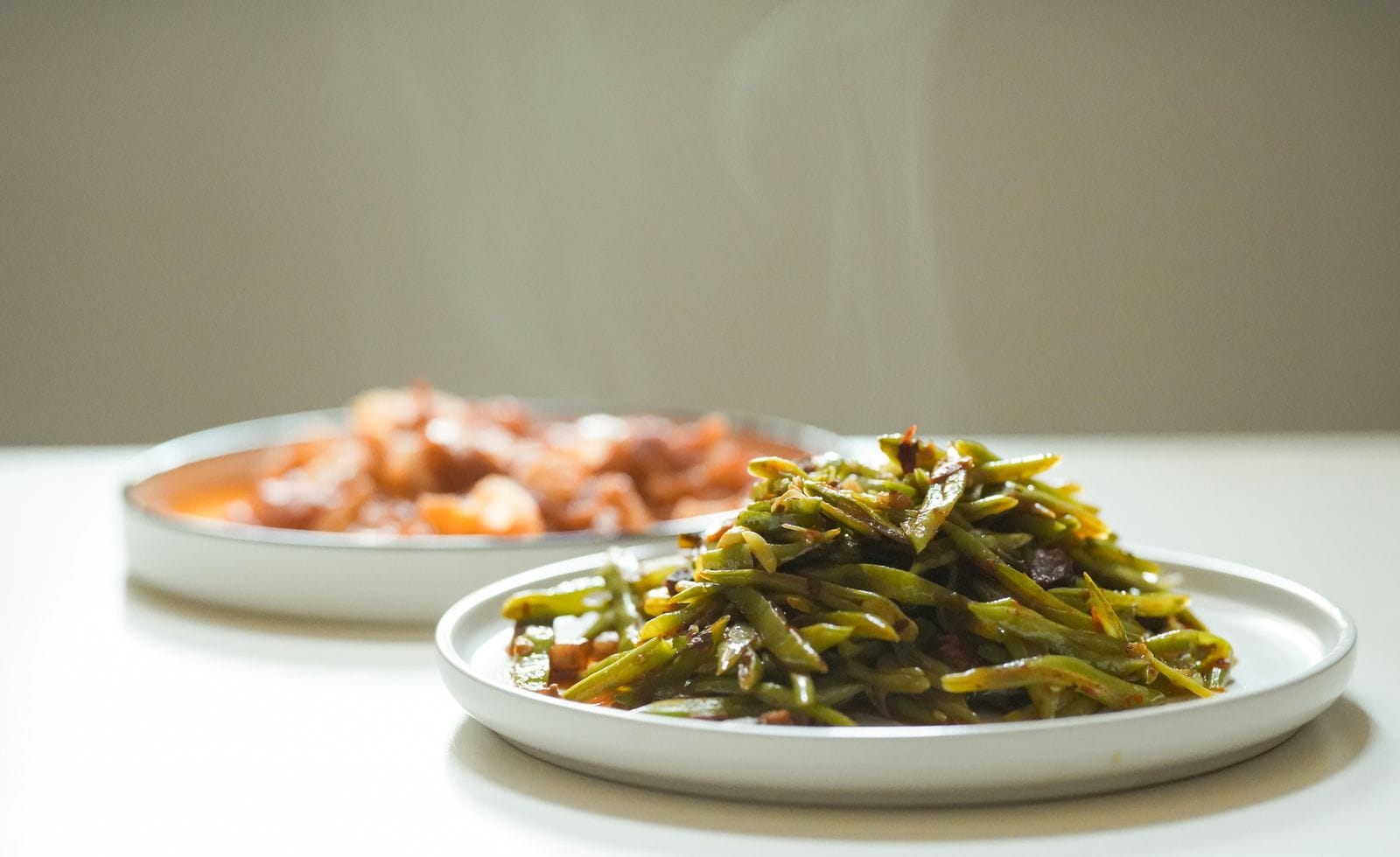 A white plate of cooked green beans with steam rising from them, a dish of stew out of focus behind