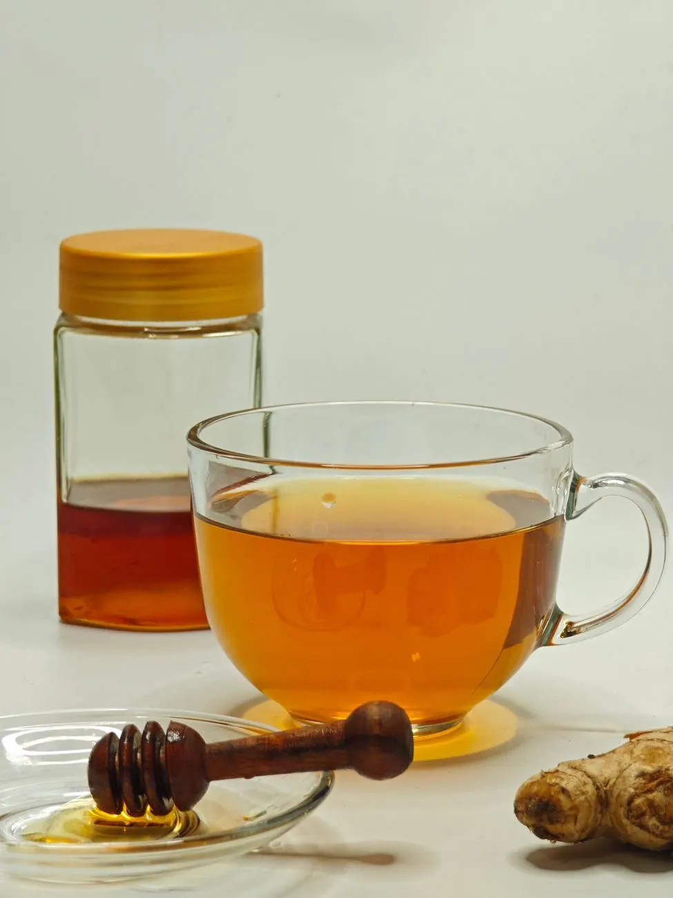 Glass cup of amber ginger tea beside a honey jar, a wooden honey dipper and a piece of fresh ginger root