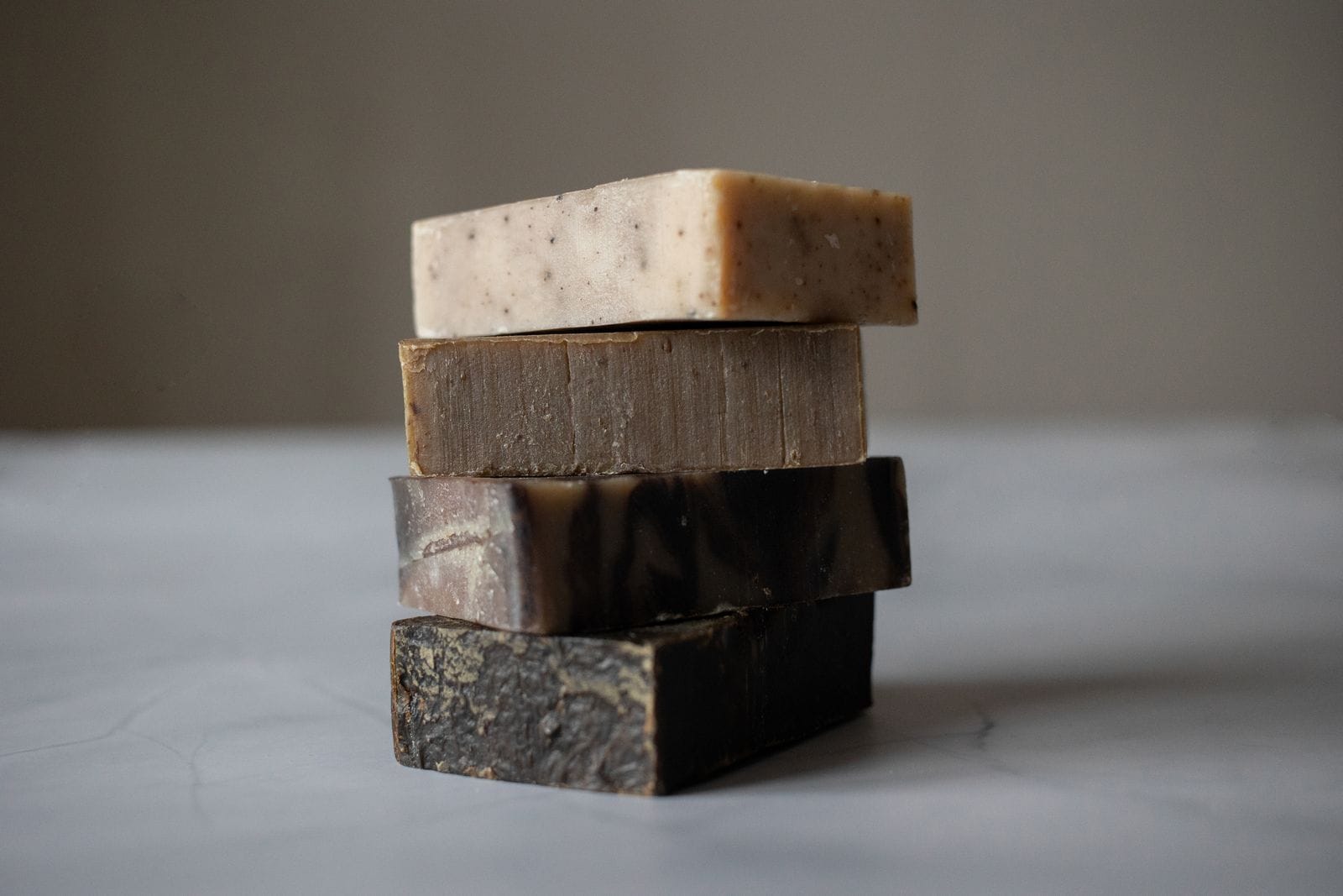 Four handmade soap bars stacked on a pale surface, ranging from speckled cream to dark brown