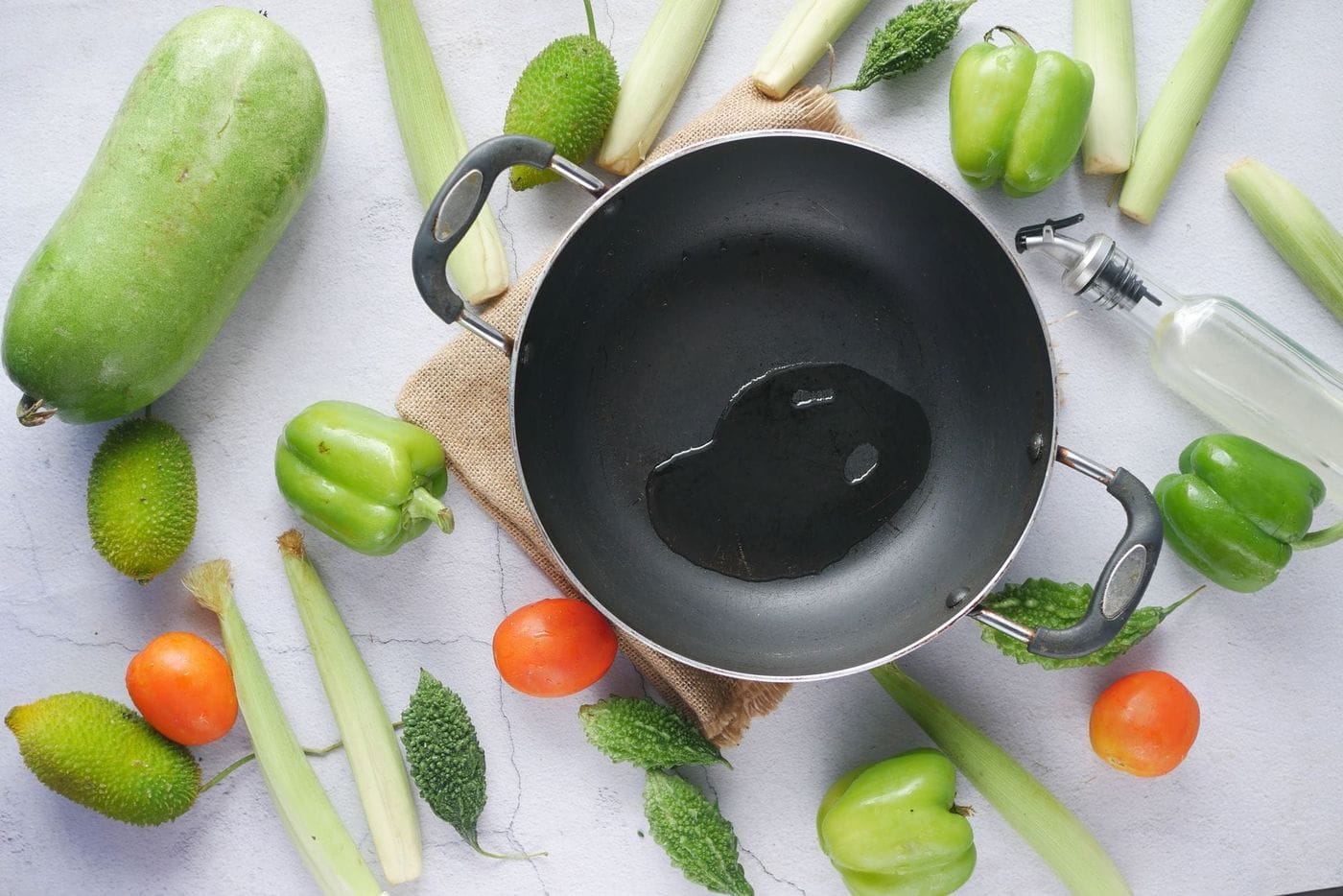 Black non-stick wok holding cooking oil, surrounded by green peppers, gourds, tomatoes and spring onions