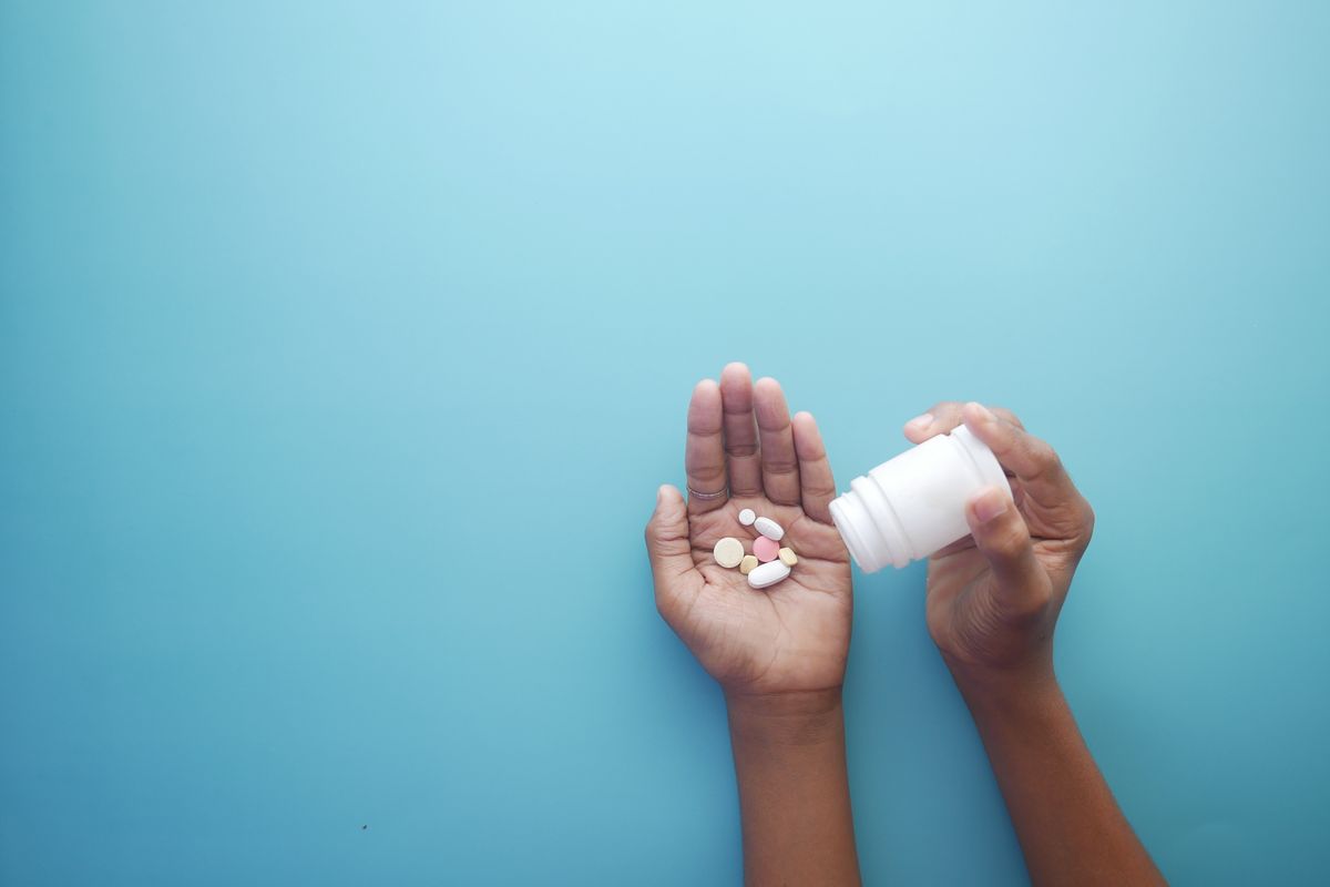 Hands tipping assorted tablets out of a plain white bottle onto an open palm against a blue background