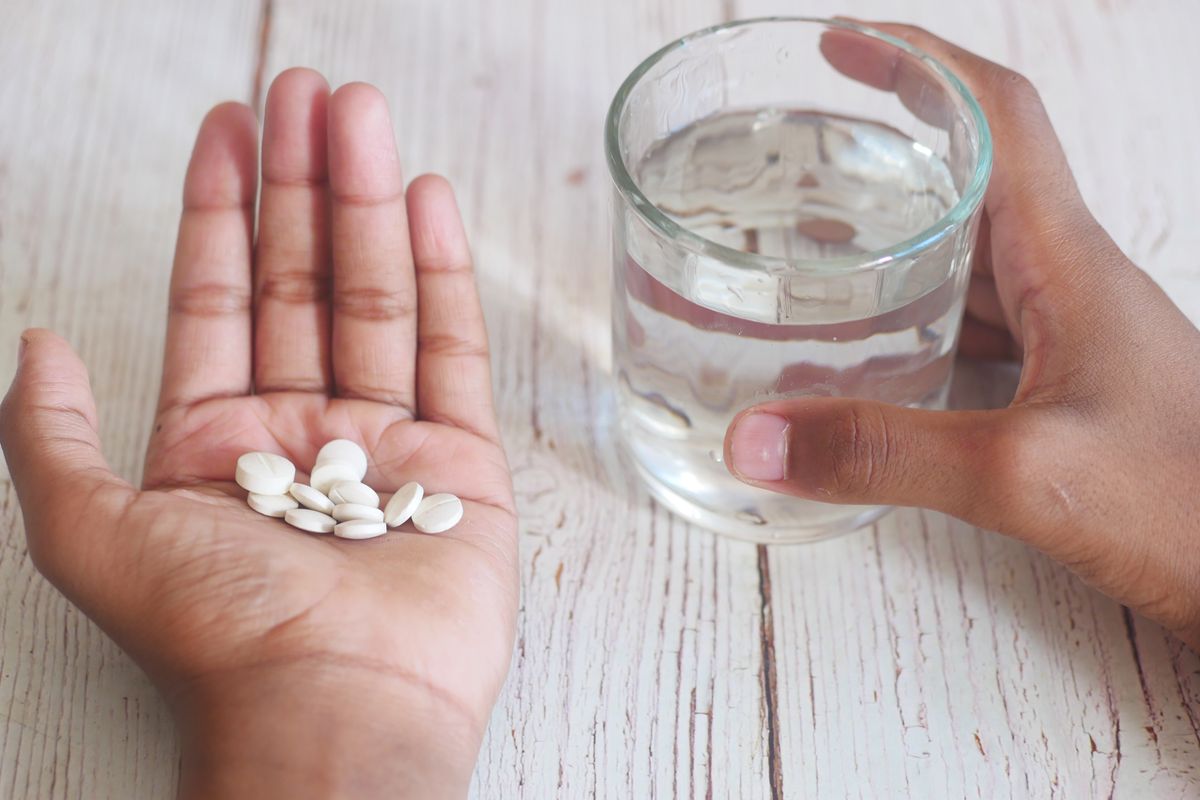 Open palm holding several white tablets beside a hand steadying a glass of water on a white wooden table