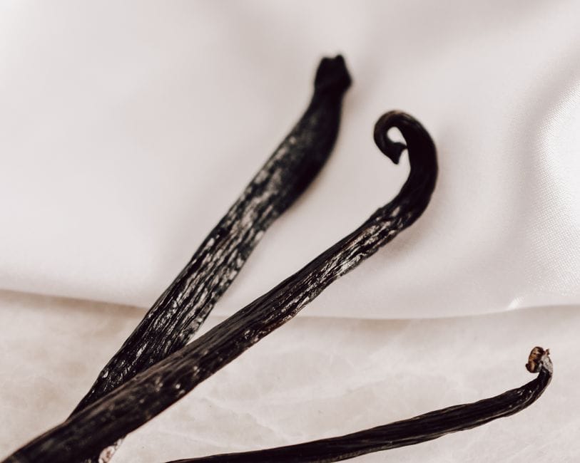 Three dark cured vanilla pods lying on a pale cream cloth, one curled at the tip