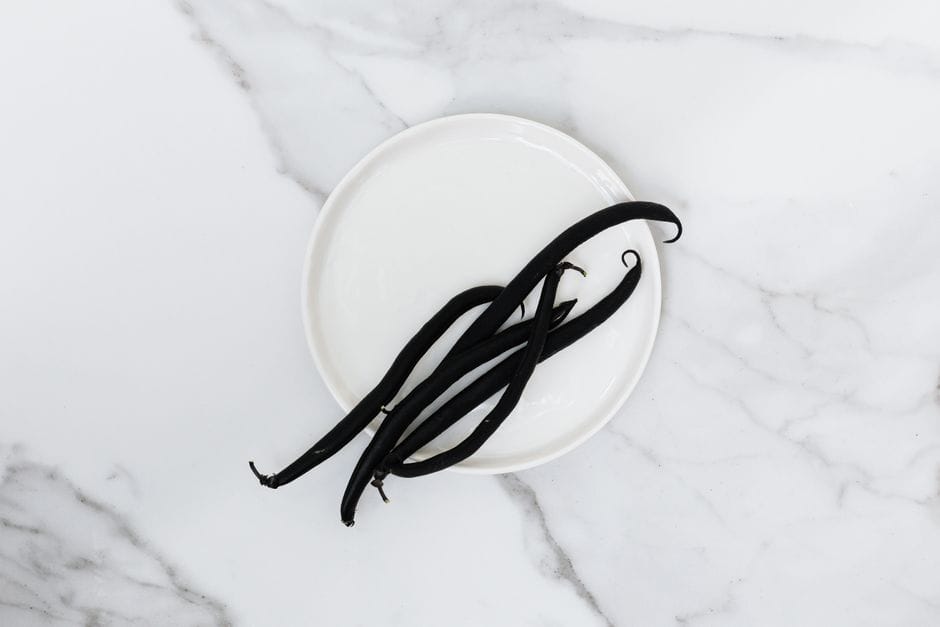 Four dark vanilla pods on a small white plate on a marble surface